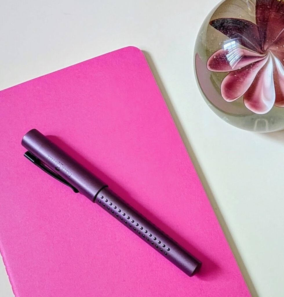 Pinkfarbenes Schreibheft mit Stift und Briefbeschwerer - Symbolbild für Kreatives Schreiben